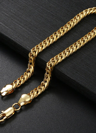 A gold-toned Cuban link chain necklace with a lobster clasp, displayed on a dark textured surface next to a black rectangular block.