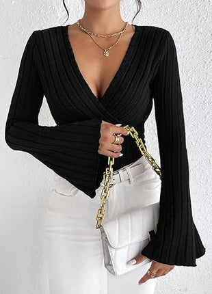 Person wearing a black long-sleeve top with a deep neckline and white pants, holding a gold chain bag against a white background.