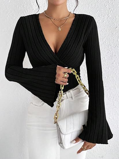 Person wearing a black long-sleeve top with a deep neckline and white pants, holding a gold chain bag against a white background.