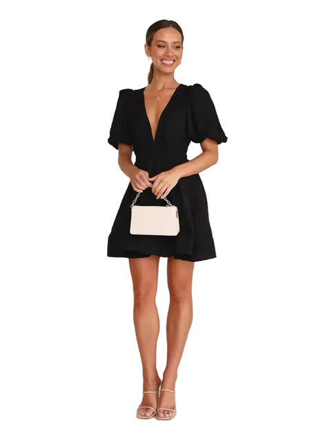 Woman in a black dress holding a white handbag against a plain background