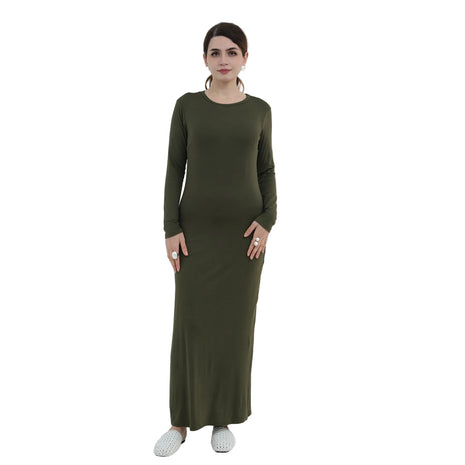 Woman wearing a long green dress standing in a minimalistic room with a white chair and decorative plant.