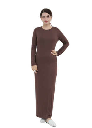 Woman wearing a brown long-sleeve dress standing in a minimalistic room with a white chair and decorative plant.