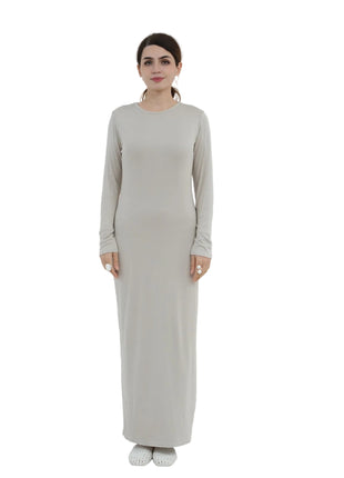 Woman wearing a long, light gray dress standing in a minimalistic room with white walls and a white chair.