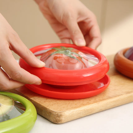 Red silicone lid being placed on a container with a tomato design, on a wooden cutting board.