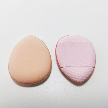Two makeup sponges, one beige and one pink with a textured edge, on a light gray background.