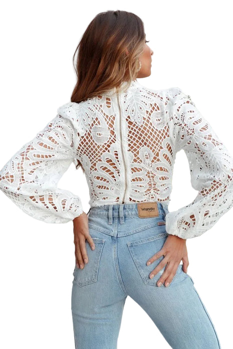 Woman wearing a white lace top and light blue jeans with a plant in the background