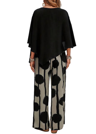 Woman wearing a black top and patterned pants standing in front of a white brick wall.