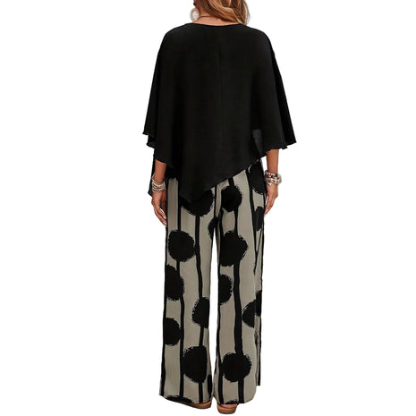 Woman wearing a black top and patterned pants standing in front of a white brick wall.