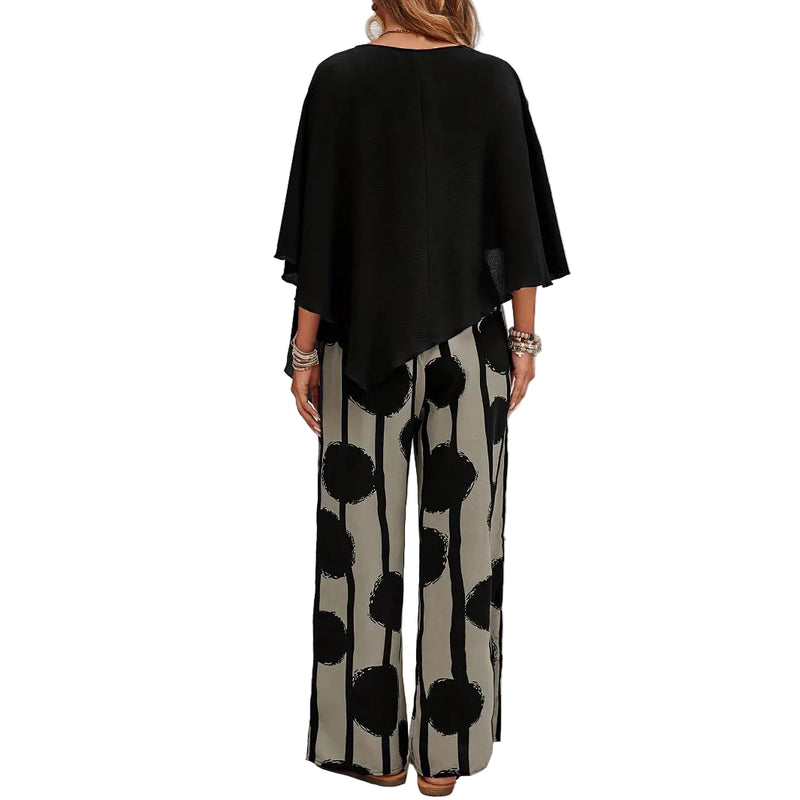 Woman wearing a black top and patterned pants standing in front of a white brick wall.