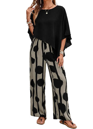 Woman wearing a black top and patterned pants standing against a white brick wall.